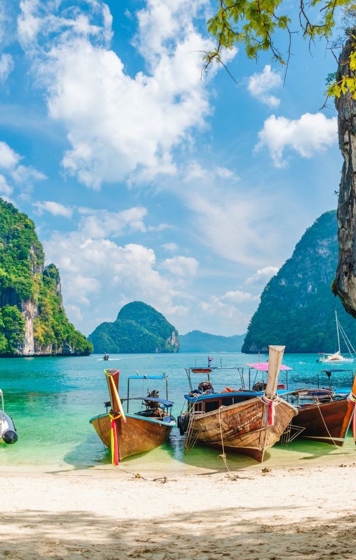 Traditional longtail boats and a motorboat docked on a sandy beach with turquoise water, surrounded by lush green cliffs under a bright blue sky.