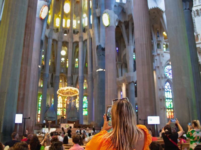 A woman in an orange dress takes a photo inside a cathedral with tall columns and colorful stained glass windows.