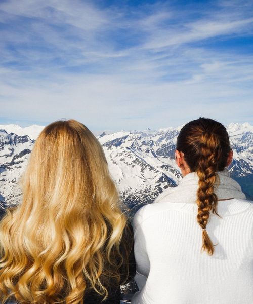 Two women with long hair, one curly blond and one brown braided, facing a snowy mountain landscape under a blue sky.
