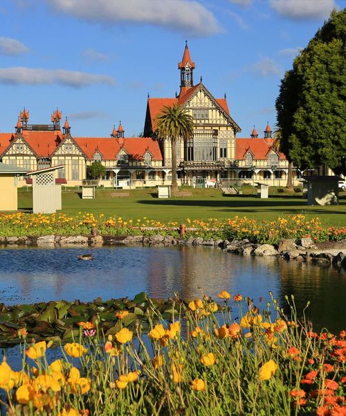 Historic building with red roofs and timber framing, surrounded by gardens, a pond with lilies, and vibrant flowers under a blue sky.