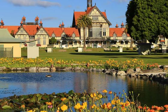 Historic building with red roofs and timber framing, surrounded by gardens, a pond with lilies, and vibrant flowers under a blue sky.