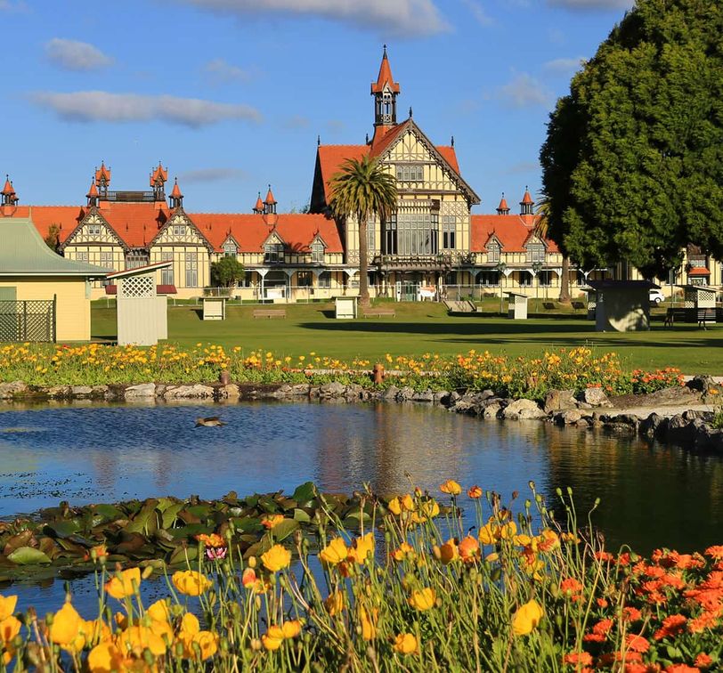 Historic building with red roofs and timber framing, surrounded by gardens, a pond with lilies, and vibrant flowers under a blue sky.