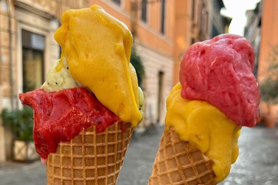 Two ice cream cones with vibrant scoops of yellow, red, and pink gelato against a background of an Italian street.