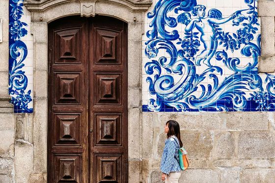 Woman walking past a large wooden door with ornate blue and white tile artwork depicting a figure and waves on the wall.