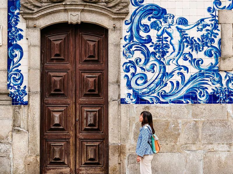 Woman walking past a large wooden door with ornate blue and white tile artwork depicting a figure and waves on the wall.