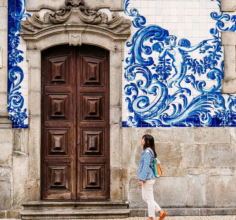 Woman walking past a large wooden door with ornate blue and white tile artwork depicting a figure and waves on the wall.