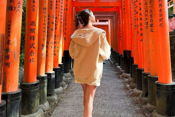 A person in a beige hoodie walks through a tunnel of red torii gates with black inscriptions, capturing a serene moment in a Japanese shrine.