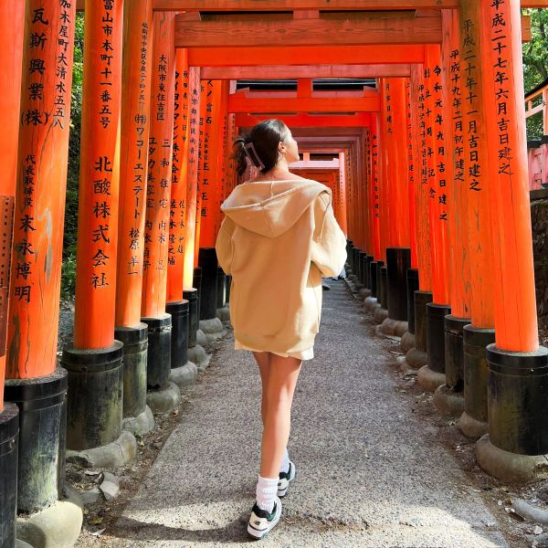 A person in a beige hoodie walks through a tunnel of red torii gates with black inscriptions, capturing a serene moment in a Japanese shrine.