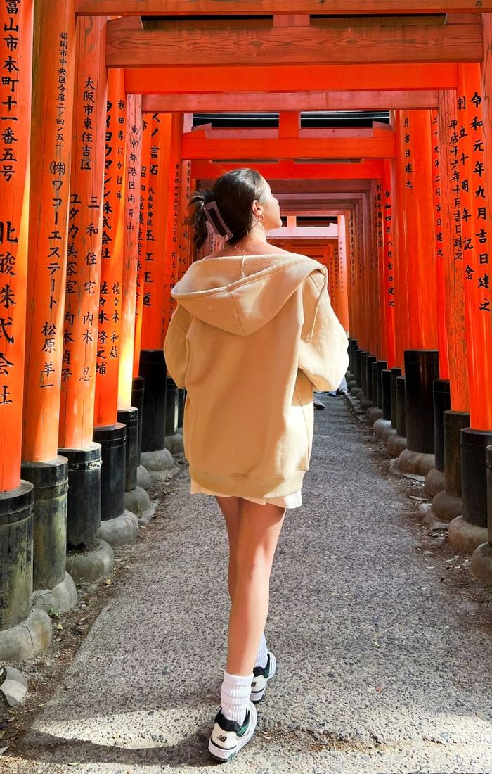 A person in a beige hoodie walks through a tunnel of red torii gates with black inscriptions, capturing a serene moment in a Japanese shrine.