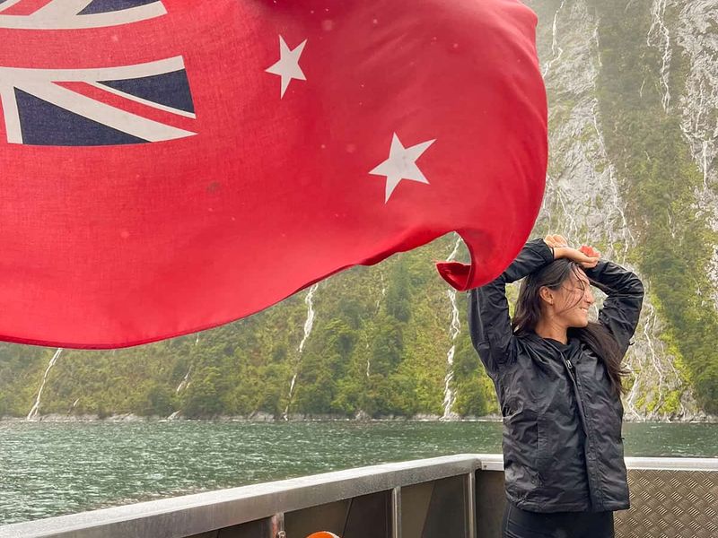 A person in a black jacket stands on a boat, looking out at a fjord, with a red flag waving nearby.