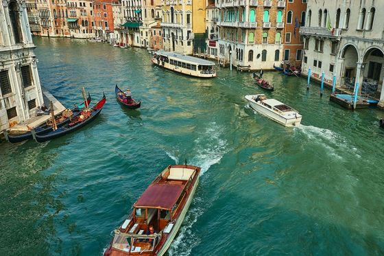 Boats and gondolas navigate a vibrant canal lined with historic buildings in Venice, featuring clear blue skies and bustling waterways.