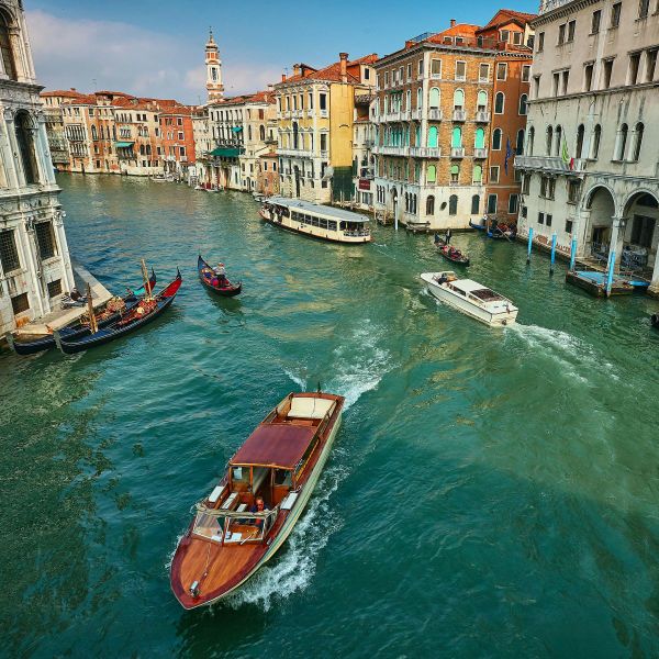 Boats and gondolas navigate a vibrant canal lined with historic buildings in Venice, featuring clear blue skies and bustling waterways.