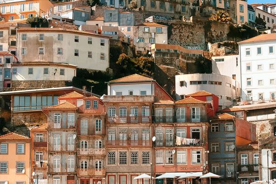Colorful hillside buildings in Porto, Portugal, with a mix of architectural styles. People gather at cafes below under umbrellas.