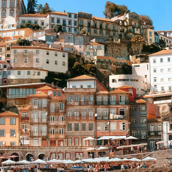 Colorful hillside buildings in Porto, Portugal, with a mix of architectural styles. People gather at cafes below under umbrellas.