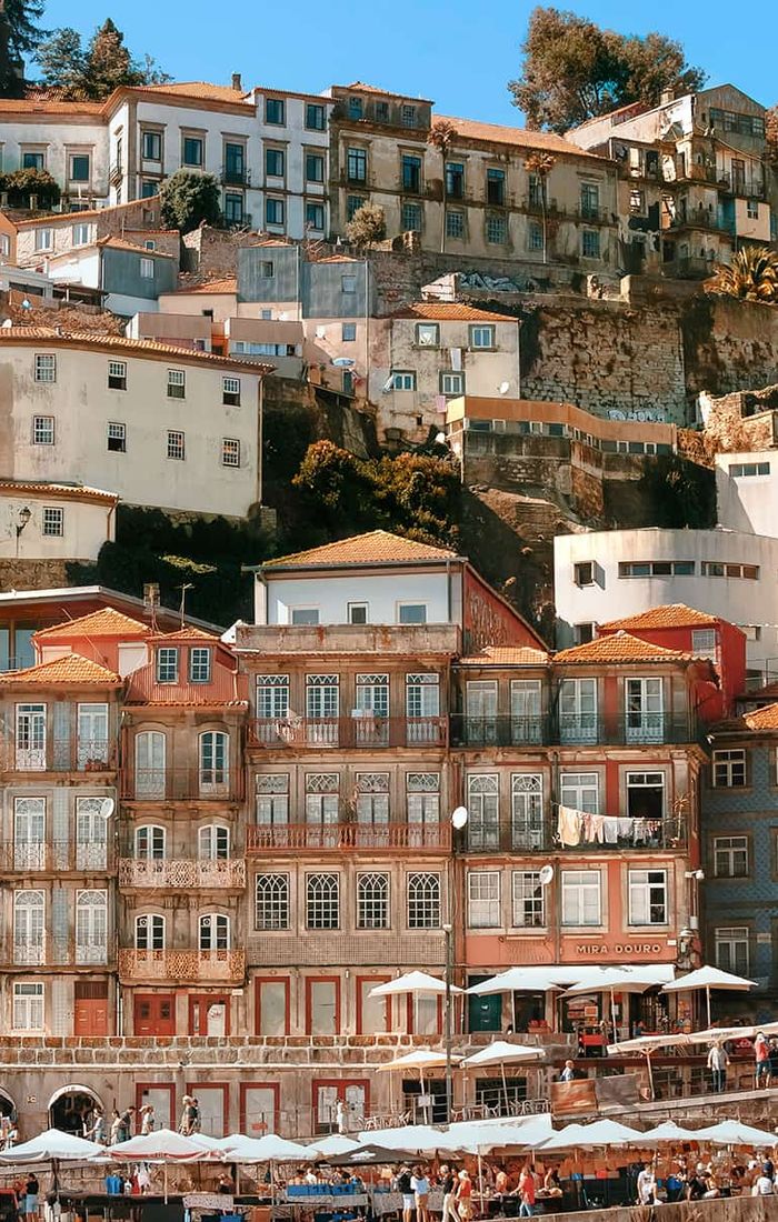 Colorful hillside buildings in Porto, Portugal, with a mix of architectural styles. People gather at cafes below under umbrellas.