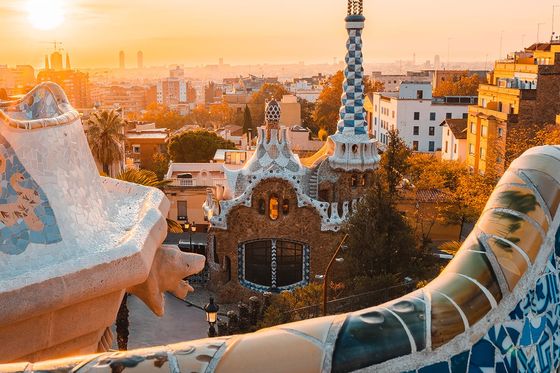 Sunrise over Park Güell in Barcelona, showcasing colorful mosaic tiles and unique architecture with a cityscape in the background.