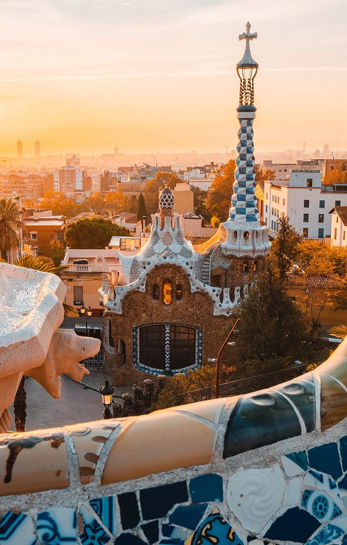 Sunrise over Park Güell in Barcelona, showcasing colorful mosaic tiles and unique architecture with a cityscape in the background.