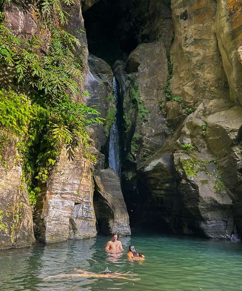 Two people swim in a clear pool at the base of a waterfall, surrounded by tall, rocky cliffs and lush green foliage.