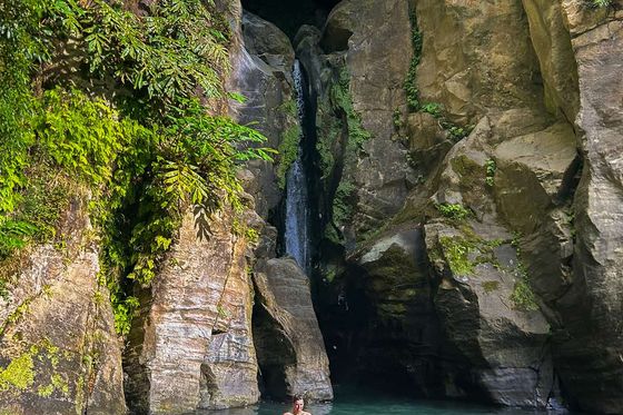 Two people swim in a clear pool at the base of a waterfall, surrounded by tall, rocky cliffs and lush green foliage.