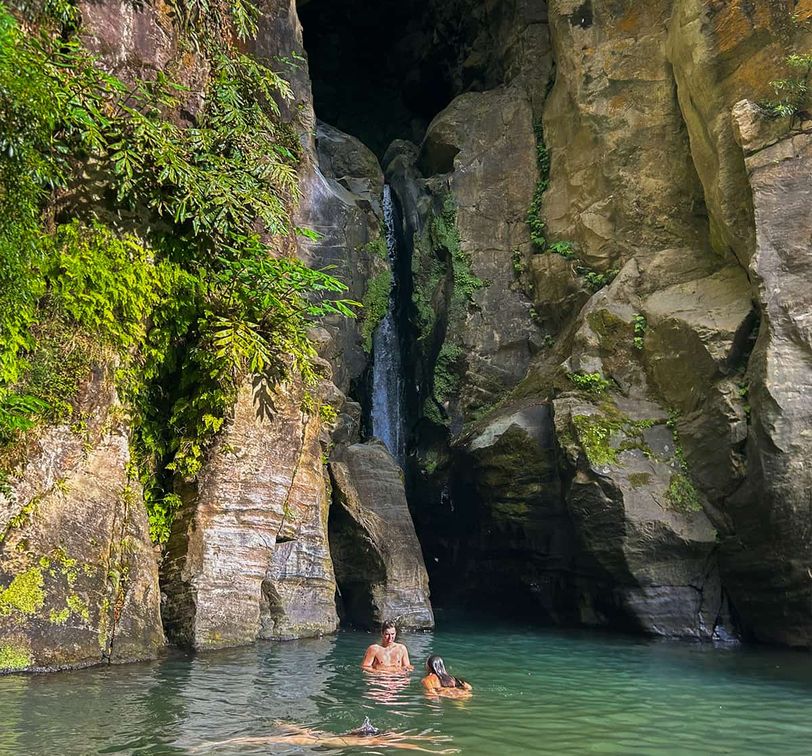 Two people swim in a clear pool at the base of a waterfall, surrounded by tall, rocky cliffs and lush green foliage.