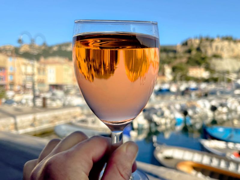 Hand holding a glass of rosé wine against a sunny marina backdrop, showcasing boats and colorful buildings.