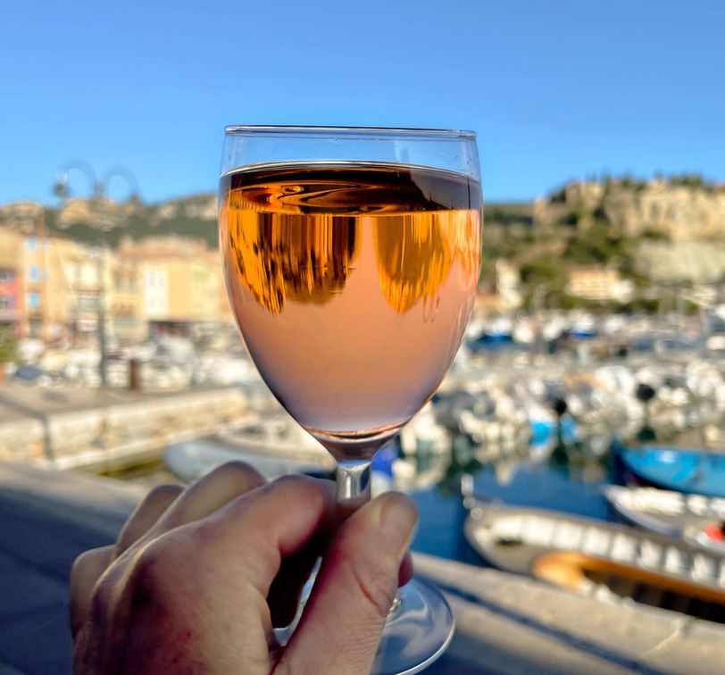 Hand holding a glass of rosé wine against a sunny marina backdrop, showcasing boats and colorful buildings.
