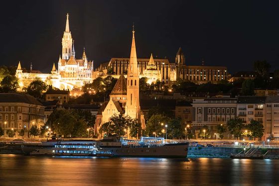 Night view of a lit-up Gothic church and buildings along a riverbank, with reflections shimmering on the water.