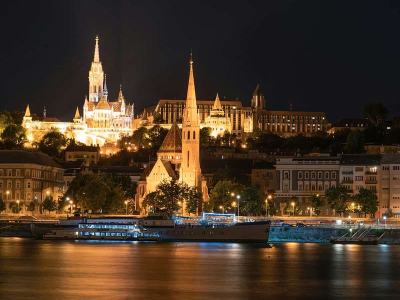 Night view of a lit-up Gothic church and buildings along a riverbank, with reflections shimmering on the water.