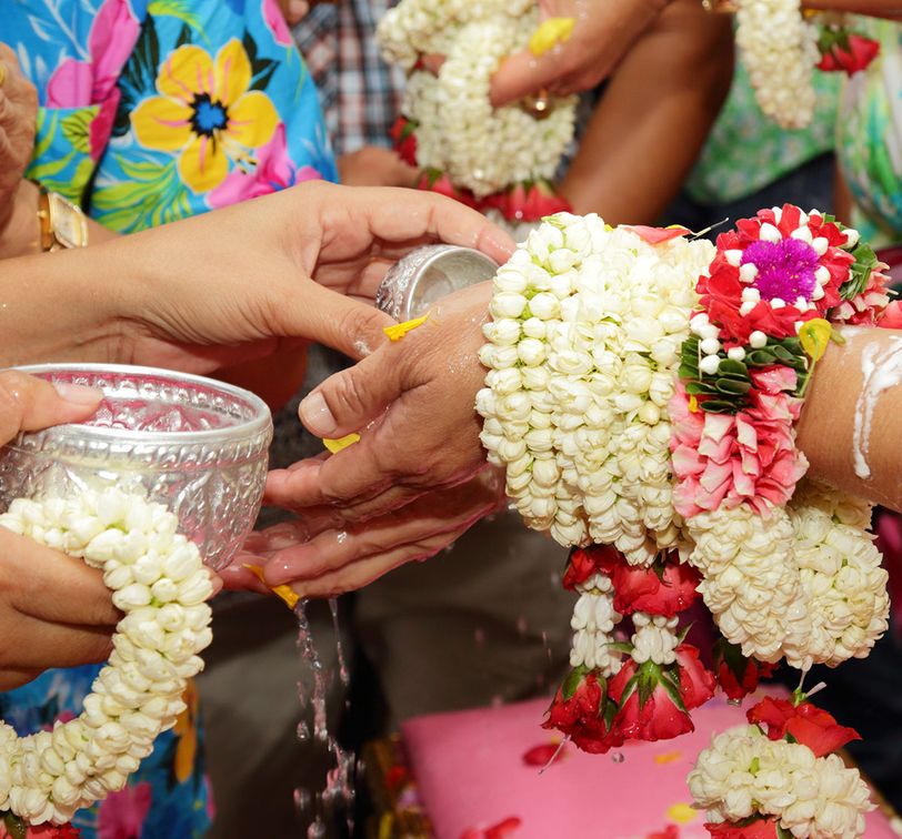 Hands with floral garlands pour water from silver bowls in a traditional ceremony, surrounded by colorful clothing and flowers.