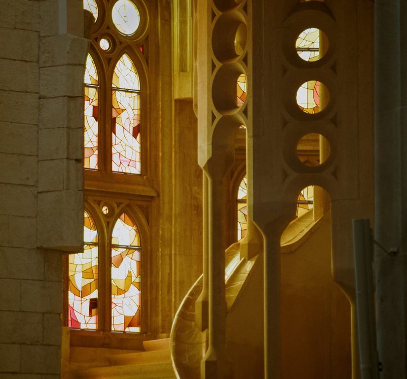 Gothic-style stained glass windows with colorful patterns light a stone staircase in a historic building interior.