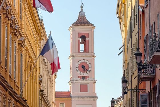 A clock tower with a pink facade between two colorful buildings, French flags waving, under a clear blue sky.