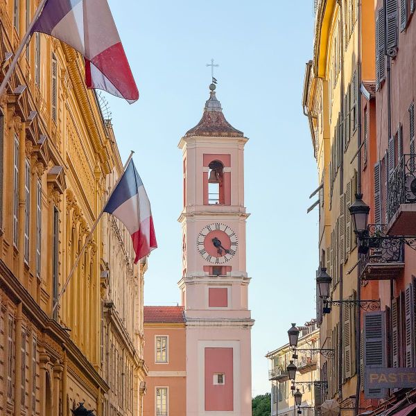 A clock tower with a pink facade between two colorful buildings, French flags waving, under a clear blue sky.