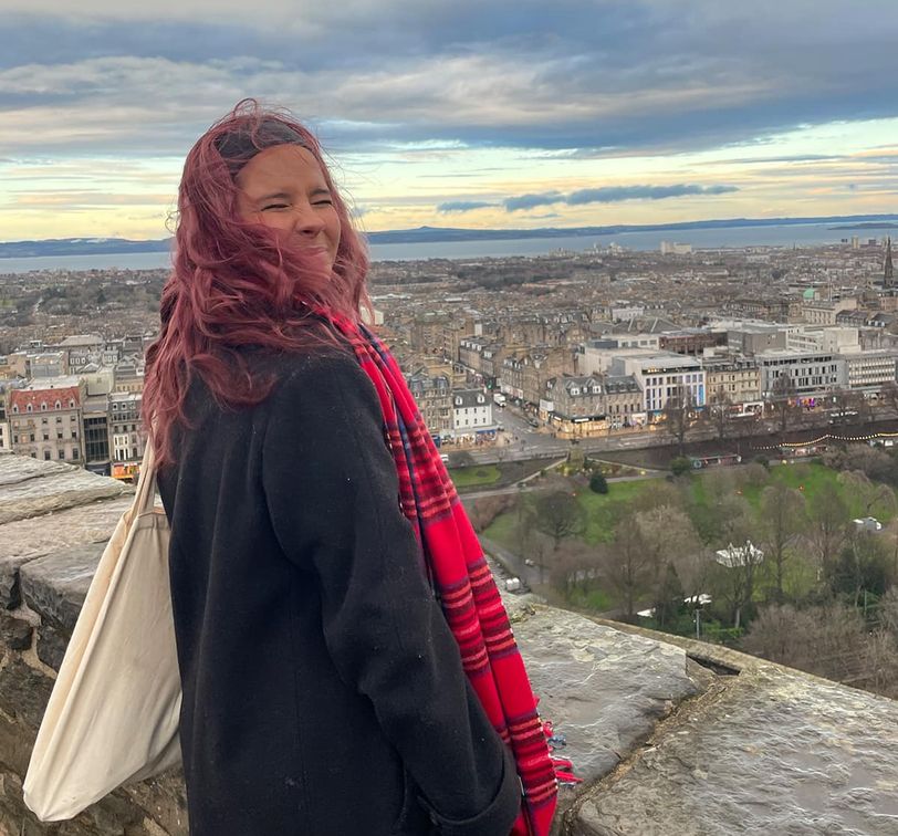 A person with red hair stands on a stone wall, overlooking a cityscape under a cloudy sky, wearing a black coat and red scarf, smiling.