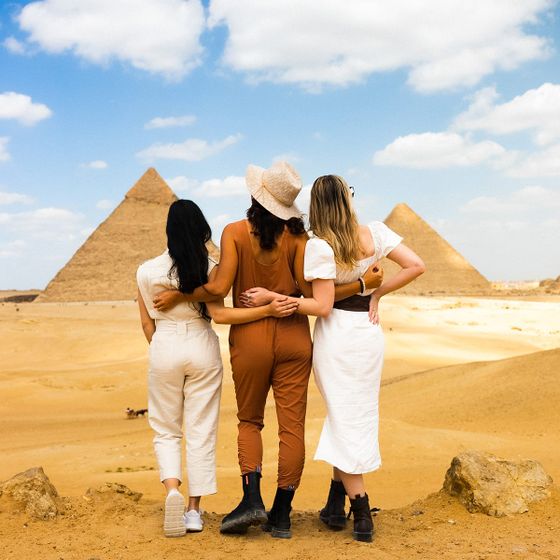Three women, arms around each other, facing Egypt's pyramids on a sunny day, with sandy desert and a blue sky in the background.