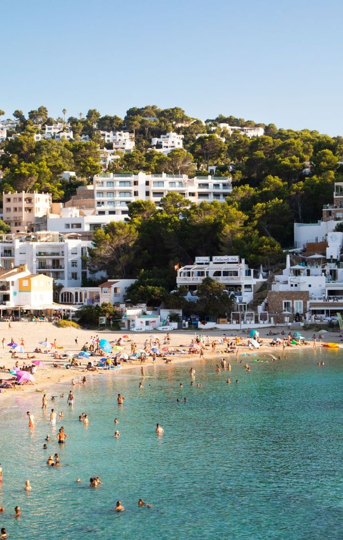Beach scene with people swimming and sunbathing; buildings and greenery in the background under a clear blue sky.