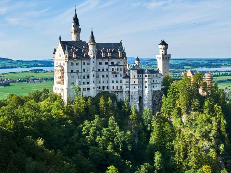 A majestic castle perched on a hilltop surrounded by lush green forests under a clear blue sky, overlooking a scenic landscape.