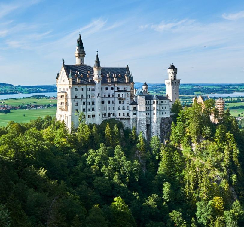 A majestic castle perched on a hilltop surrounded by lush green forests under a clear blue sky, overlooking a scenic landscape.