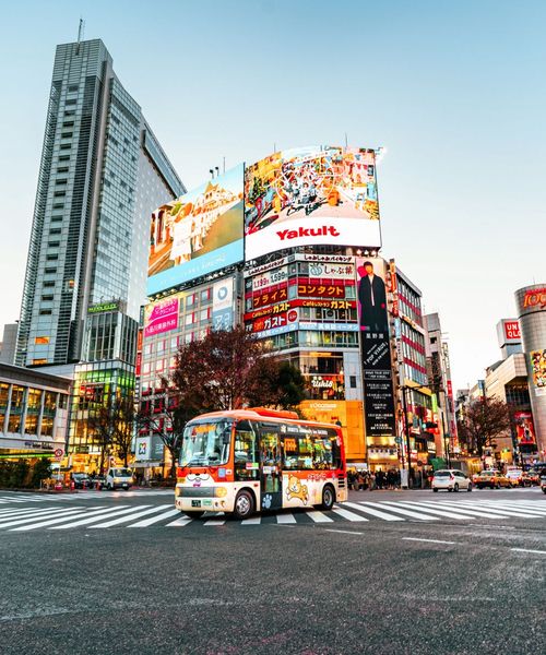 A vibrant city scene with a colorful bus on a busy street lined with tall buildings and large illuminated billboards, captured at dusk.