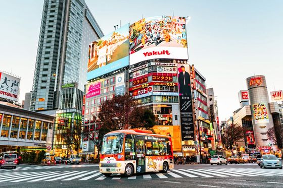 A vibrant city scene with a colorful bus on a busy street lined with tall buildings and large illuminated billboards, captured at dusk.