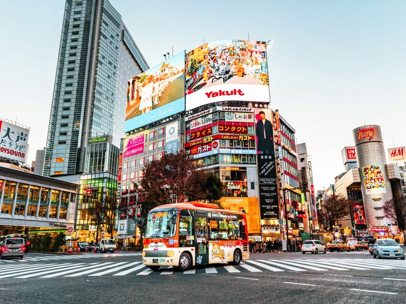 A vibrant city scene with a colorful bus on a busy street lined with tall buildings and large illuminated billboards, captured at dusk.