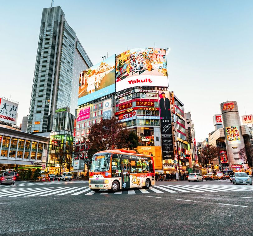 A vibrant city scene with a colorful bus on a busy street lined with tall buildings and large illuminated billboards, captured at dusk.