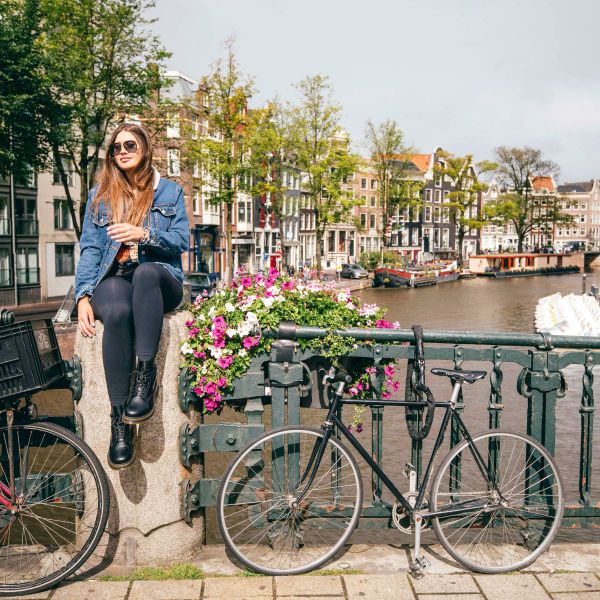 Woman in a denim jacket sits on a bridge's stone ledge beside a bicycle, overlooking a canal with colorful buildings and a boat.