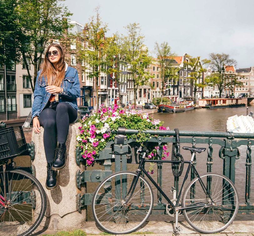 Woman in a denim jacket sits on a bridge's stone ledge beside a bicycle, overlooking a canal with colorful buildings and a boat.