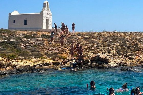 People swim and relax in clear blue waters near a rocky shore with a small white chapel on a hill under a bright blue sky.