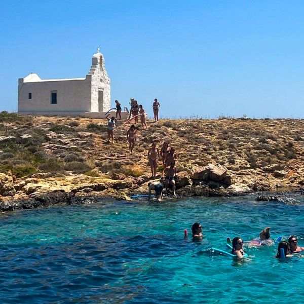 People swim and relax in clear blue waters near a rocky shore with a small white chapel on a hill under a bright blue sky.