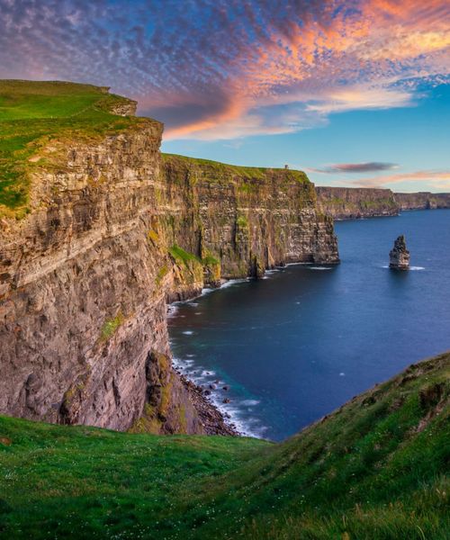 Cliffs of Moher with lush green grass, rocky cliffs, and a vast ocean under a colorful sunset sky.