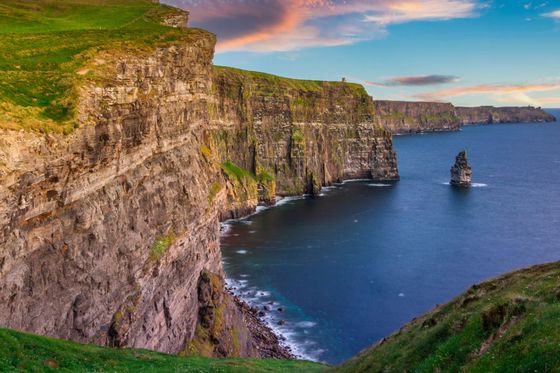 Cliffs of Moher with lush green grass, rocky cliffs, and a vast ocean under a colorful sunset sky.
