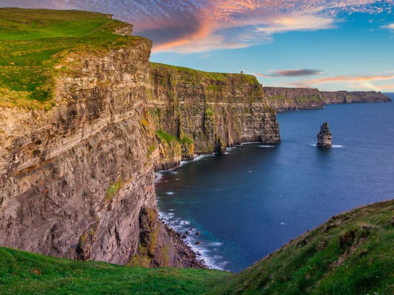 Cliffs of Moher with lush green grass, rocky cliffs, and a vast ocean under a colorful sunset sky.