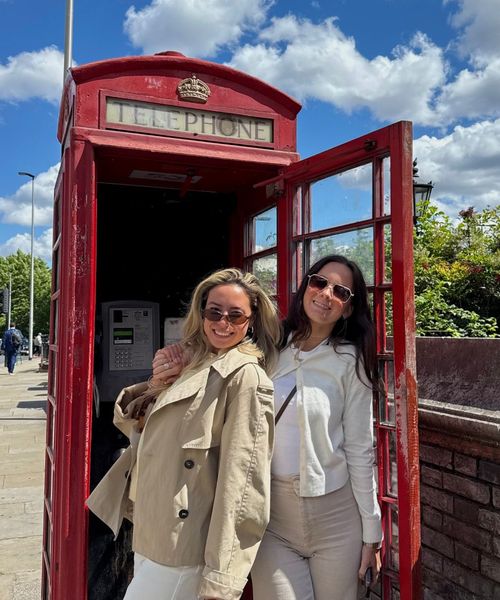 Two women smiling in front of a classic red British telephone booth on a sunny day, with blue sky and fluffy clouds in the background.