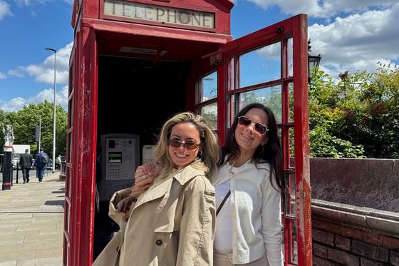 Two women smiling in front of a classic red British telephone booth on a sunny day, with blue sky and fluffy clouds in the background.